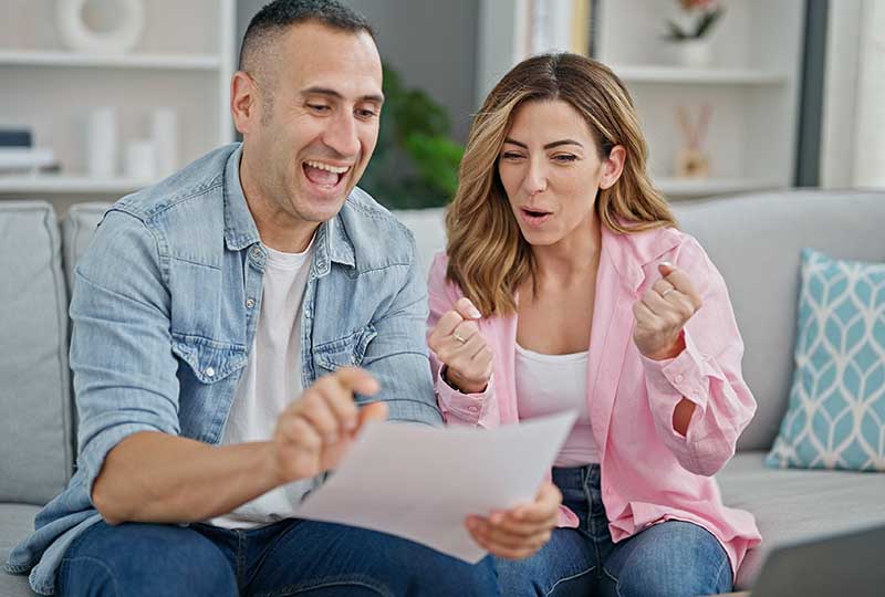 A man and woman are sitting on a couch, looking at papers with expressions of surprise or interest.